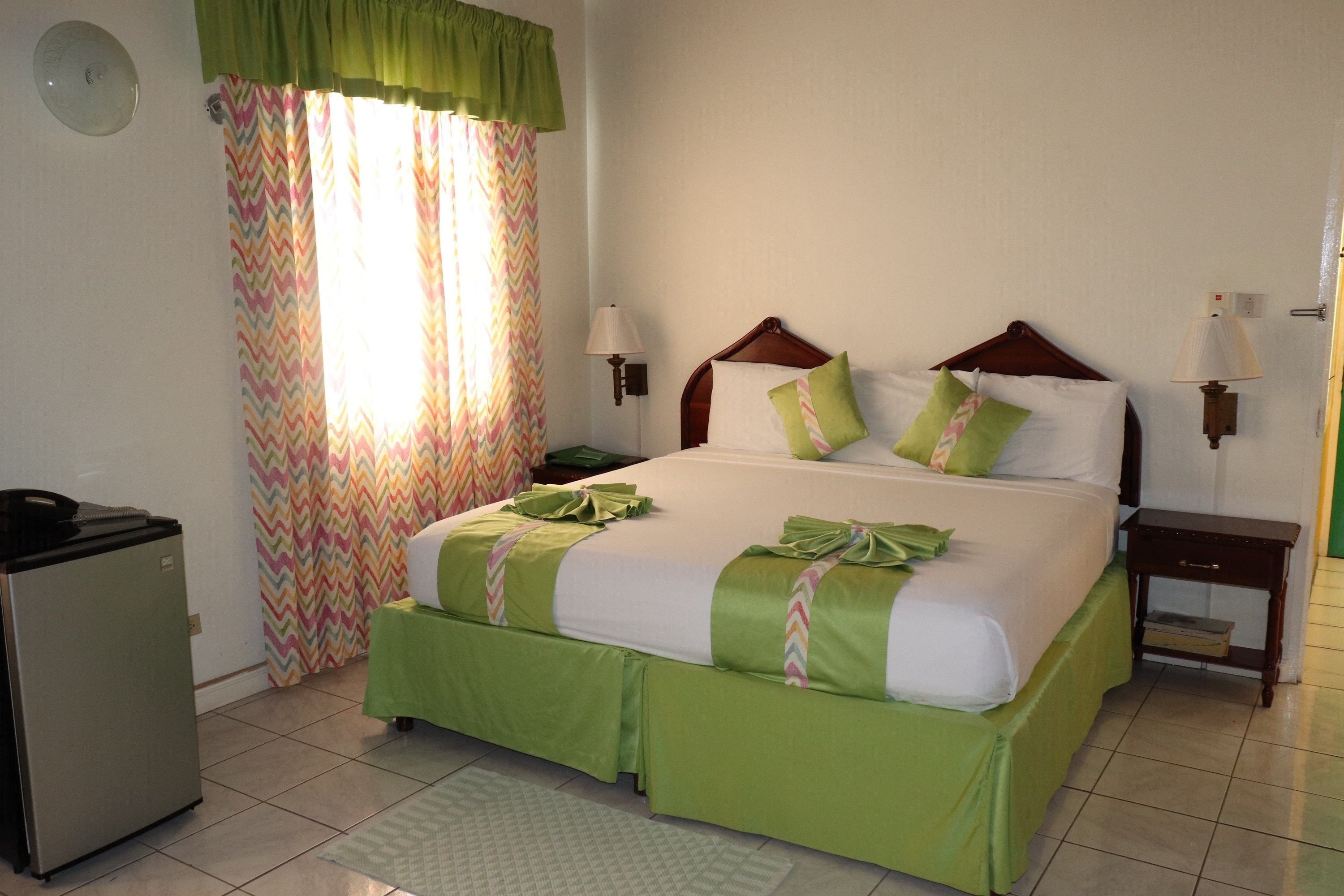 A neatly arranged bedroom with a bed, bedside lamps, a small table, and a refrigerator.