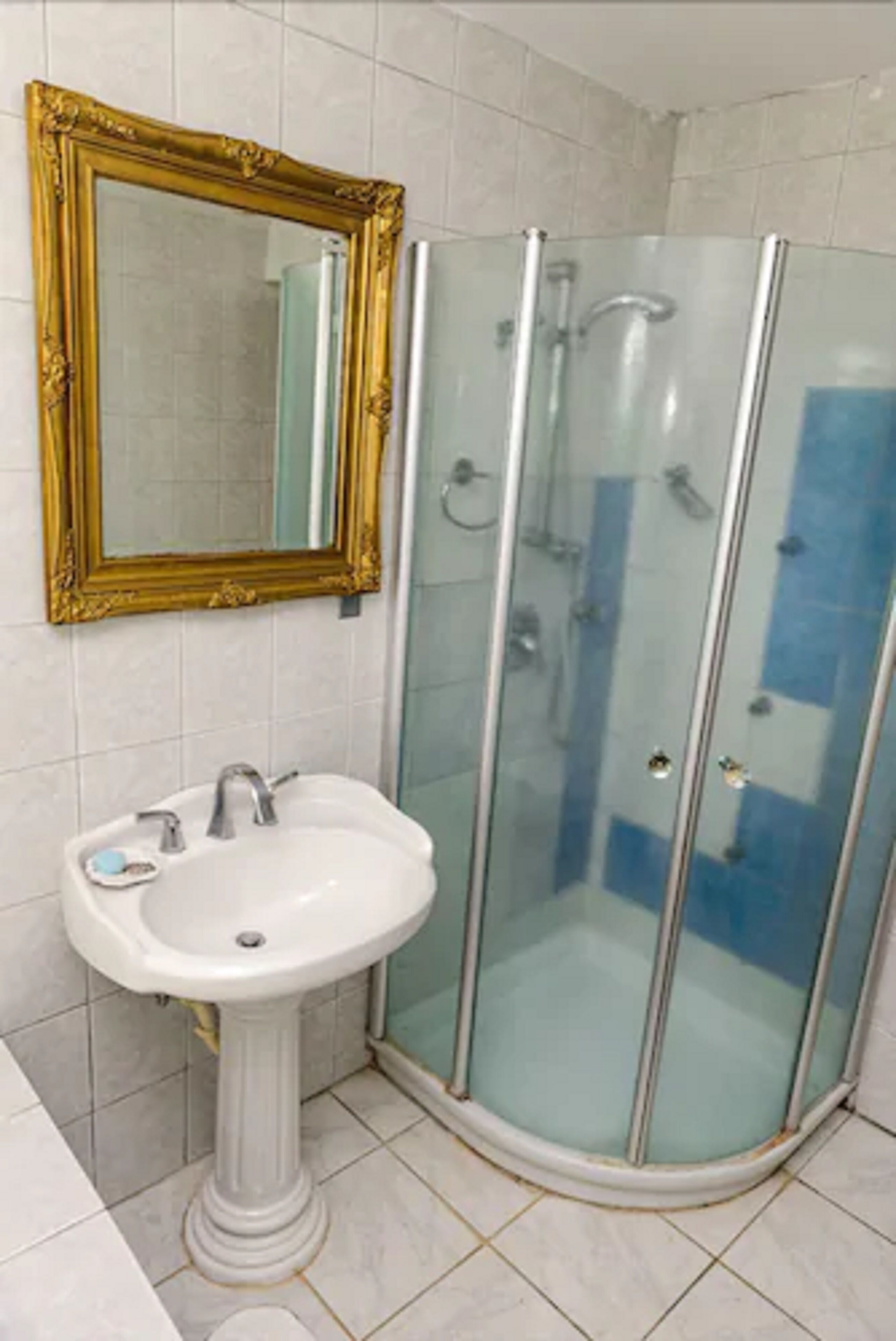 A bathroom with a pedestal sink, a gold-framed mirror, and a glass-enclosed shower.