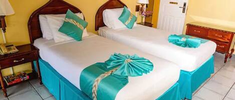 A hotel room with two beds, each with a wooden headboard, white linens, and teal bed skirts. There is a nightstand with a lamp and a small table with a book.