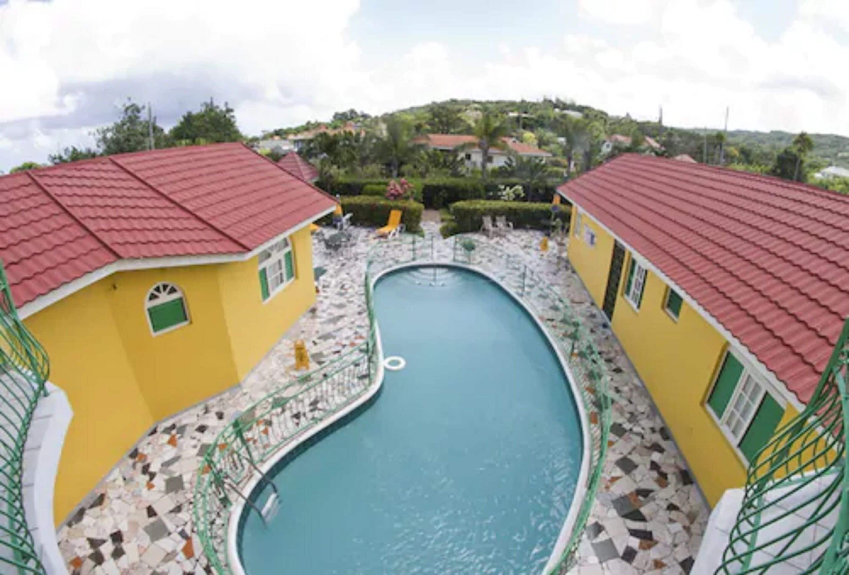A pool area with two yellow buildings, each with a red roof, surrounded by a tiled deck.