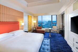 Ein Hotelzimmer mit einem großen Bett, einem Schreibtisch, einem Stuhl, einem Fernseher und Blick auf die Stadt.