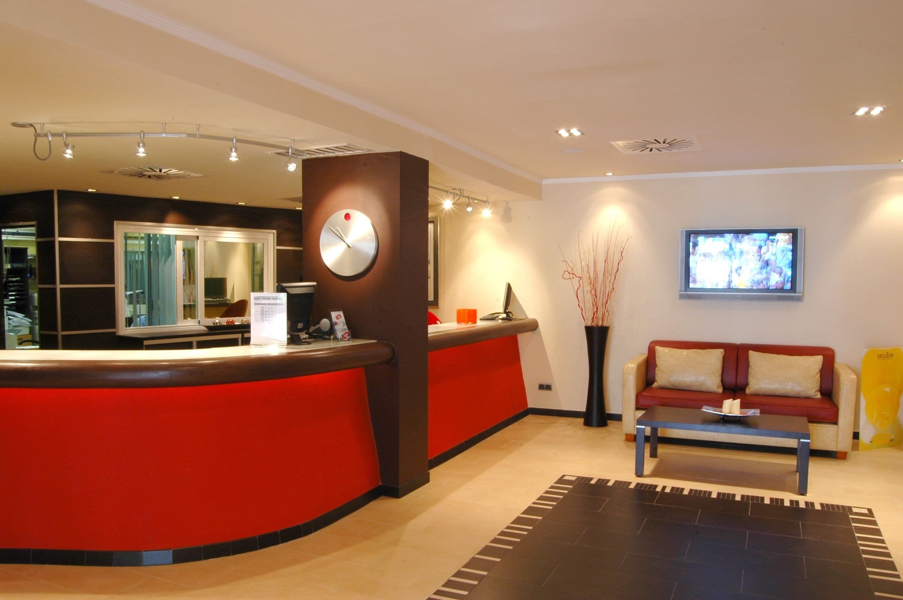 Zona de recepción con mostrador rojo, un reloj, televisor, sofá y mesa de centro.