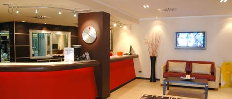 Zona de recepción con mostrador rojo, un reloj, televisor, sofá y mesa de centro.