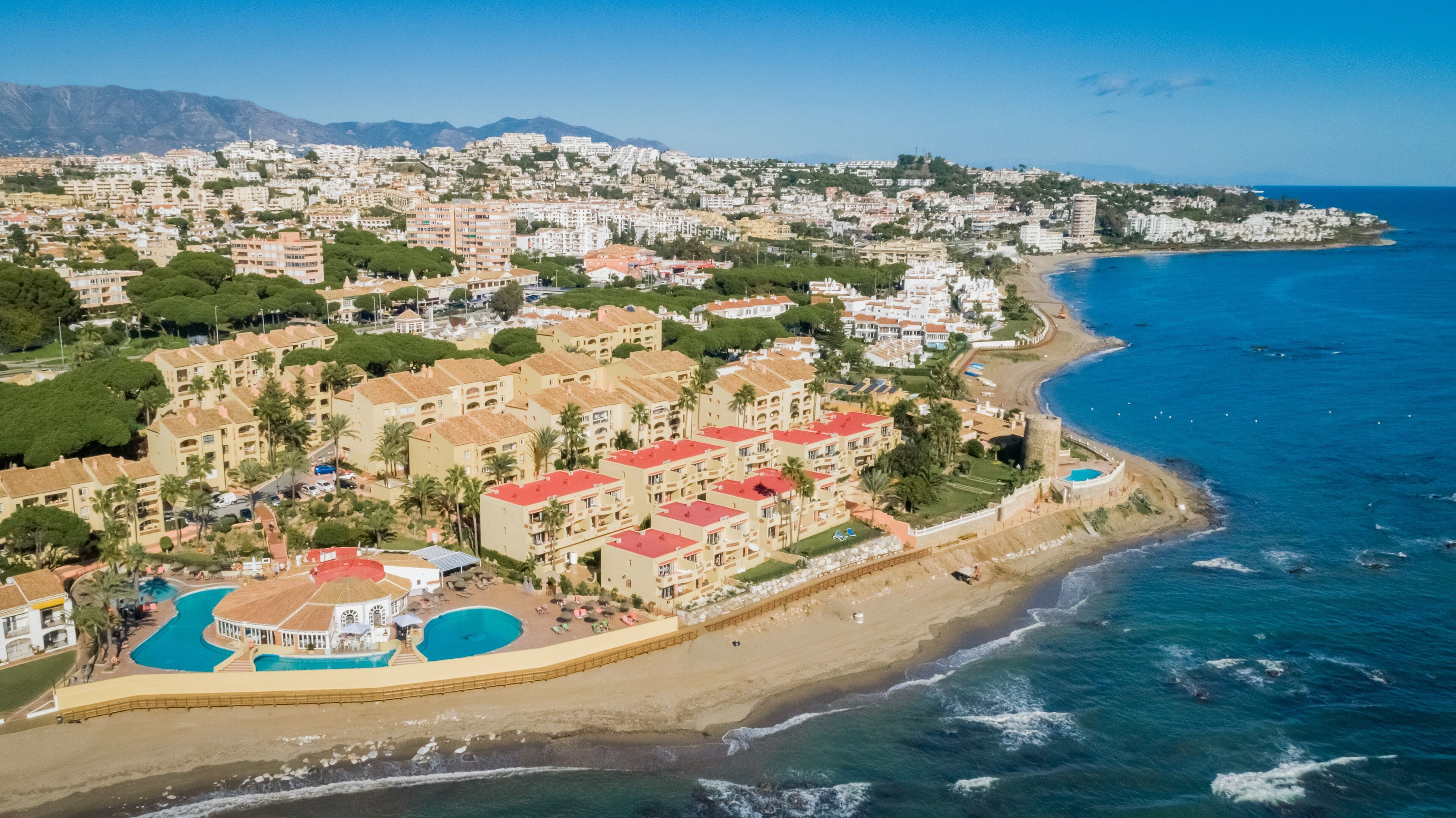Vista aérea de una zona residencial costera con playa, que incluye una piscina y una playa con sombrillas.