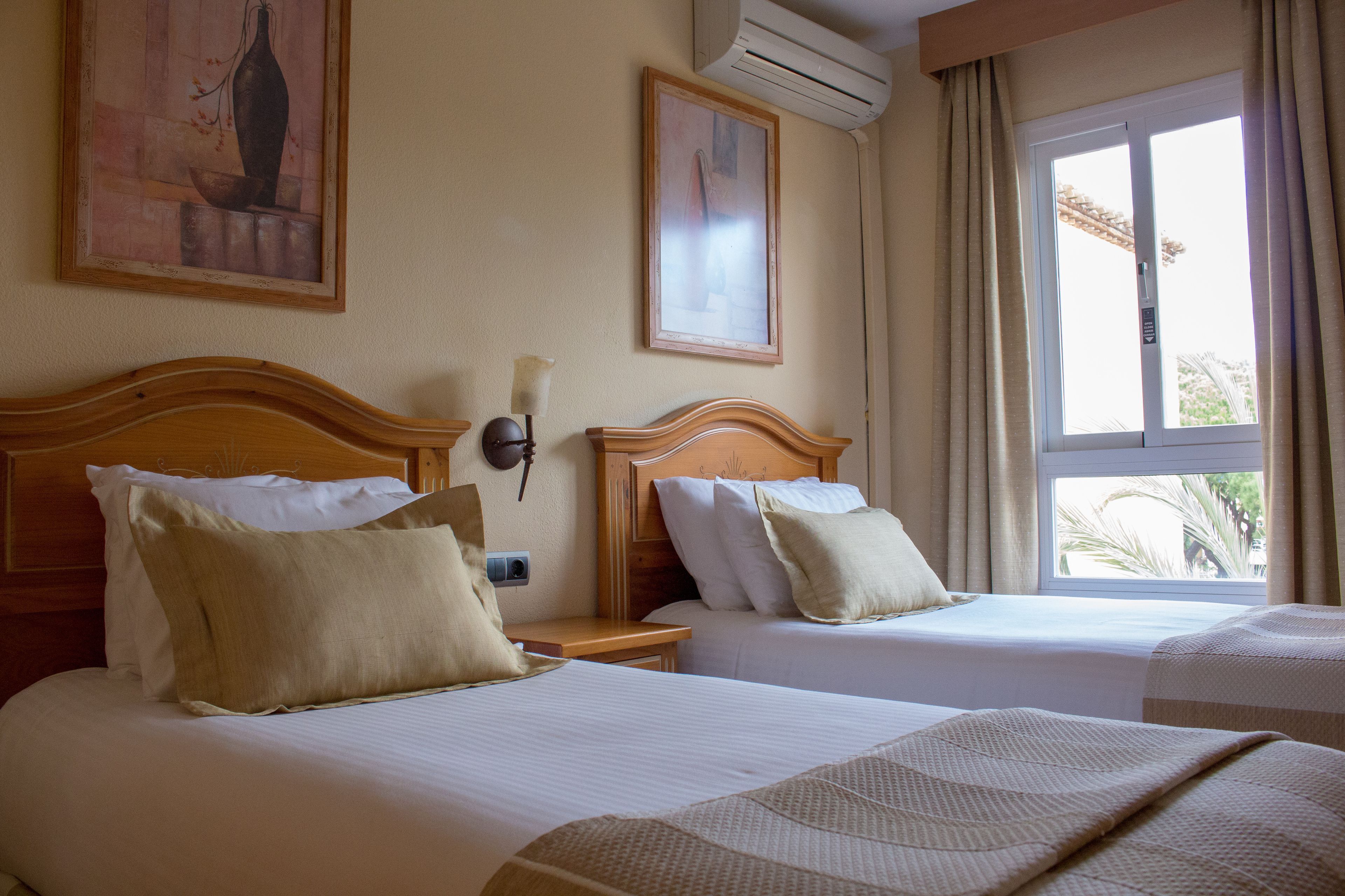 A hotel room with two beds, a nightstand, a wall-mounted lamp, and a window with curtains.