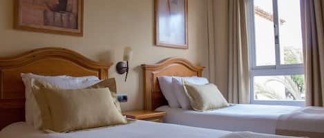 A hotel room with two beds, a nightstand, a wall-mounted lamp, and a window with curtains.