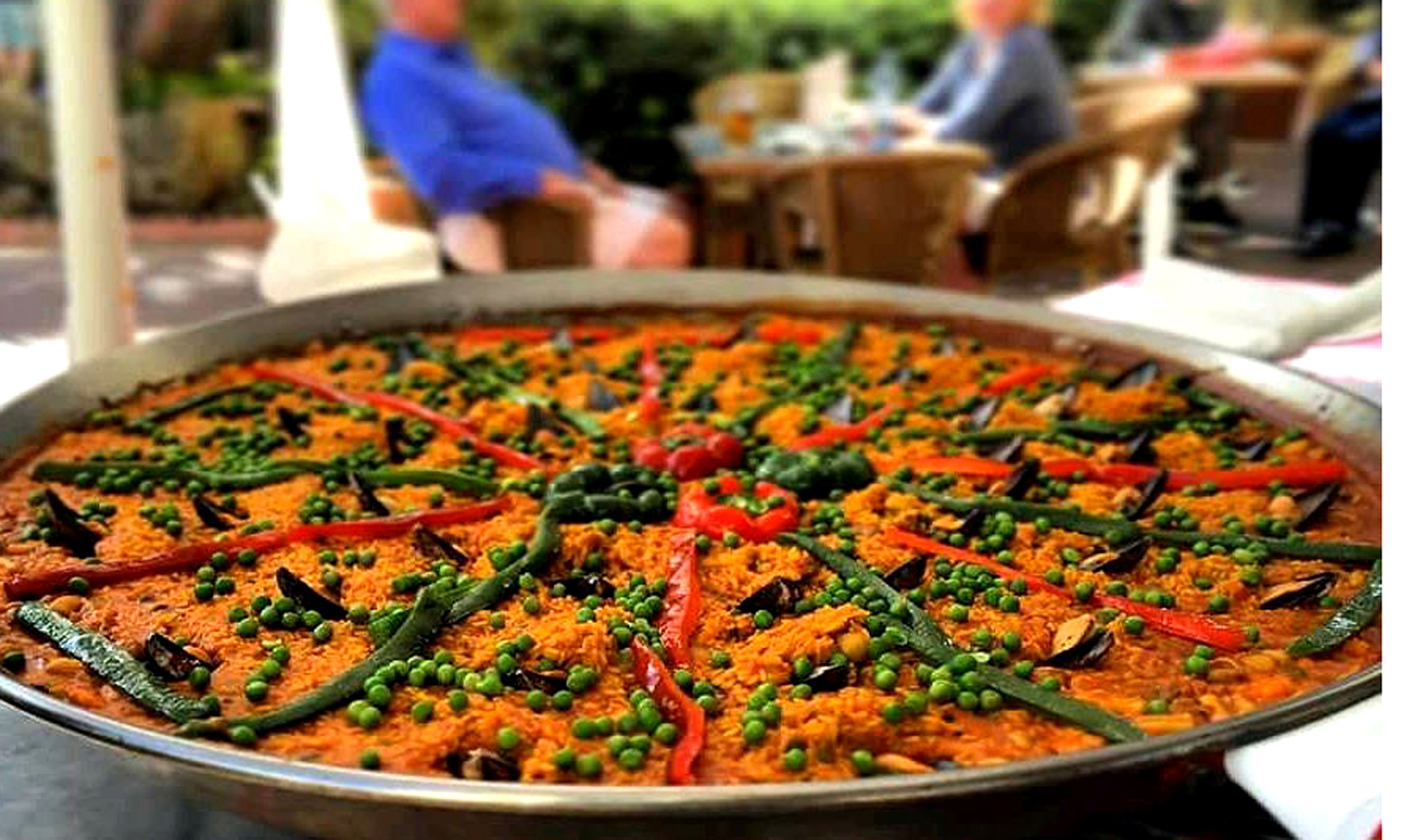 Una paella grande con verduras y mariscos, servida en una mesa al aire libre.
