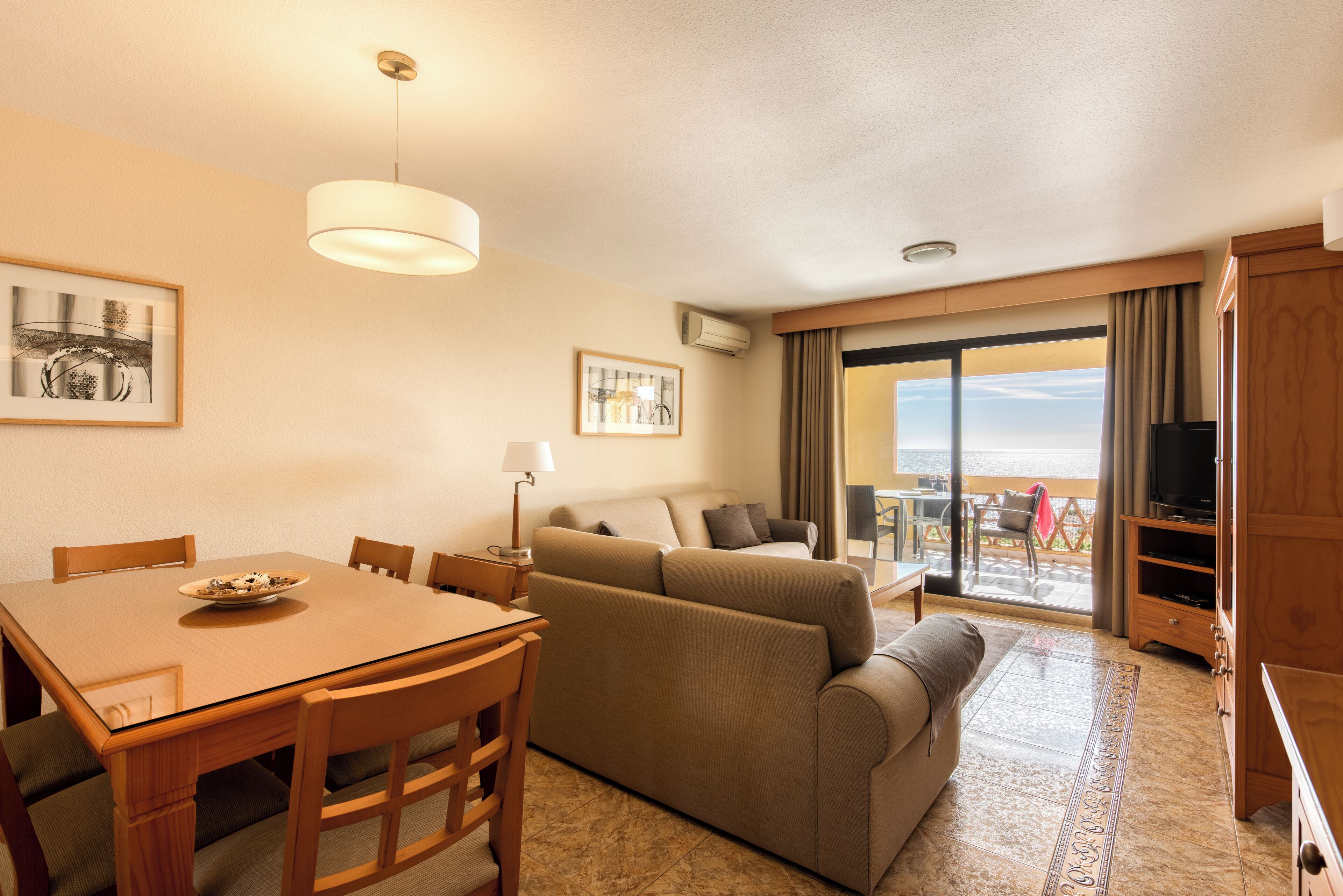 A hotel room with a dining table, chairs, a sofa, a TV, and a balcony with a view.
