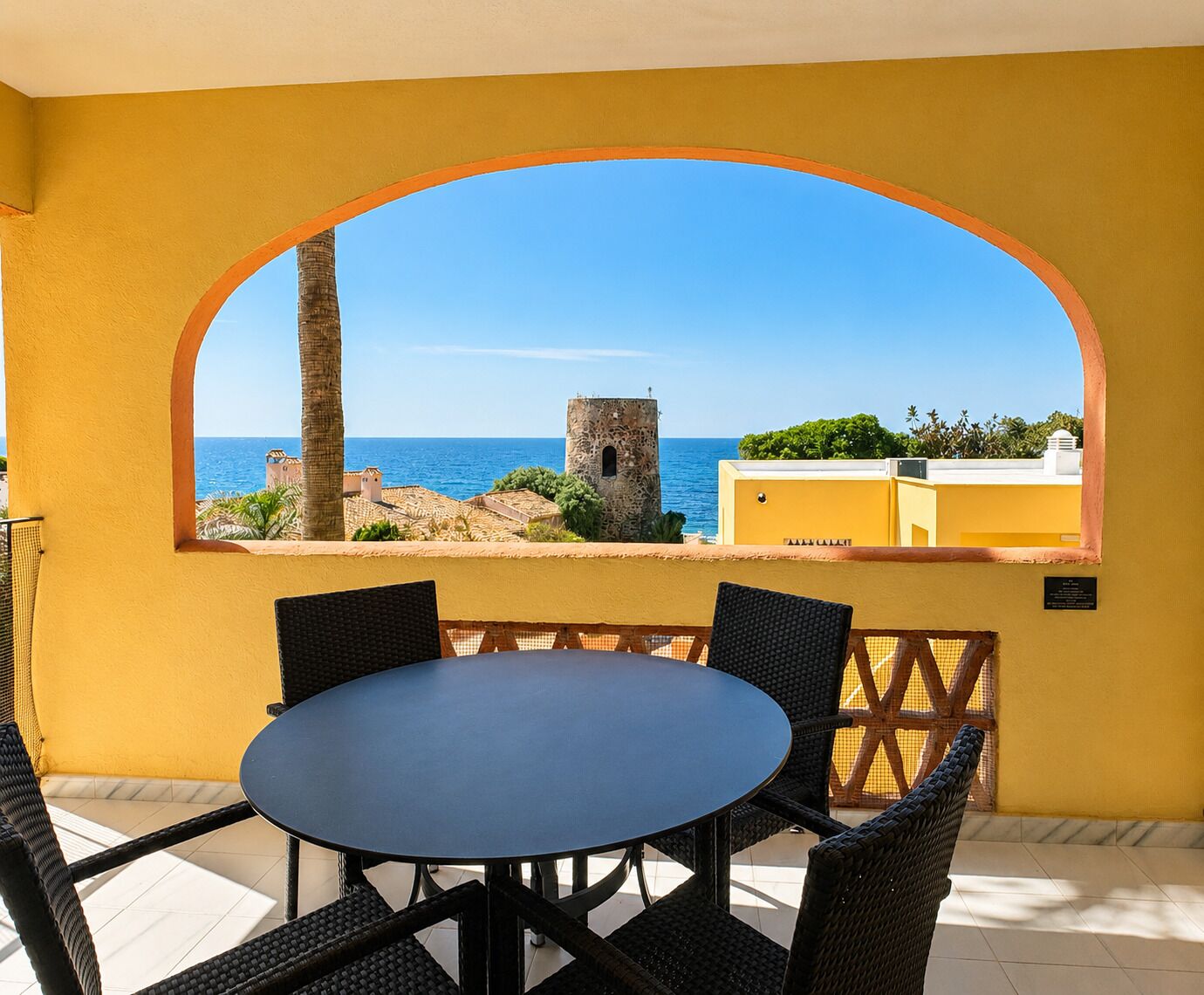 Un balcón con mesa y sillas, vista al mar y una torre al fondo.