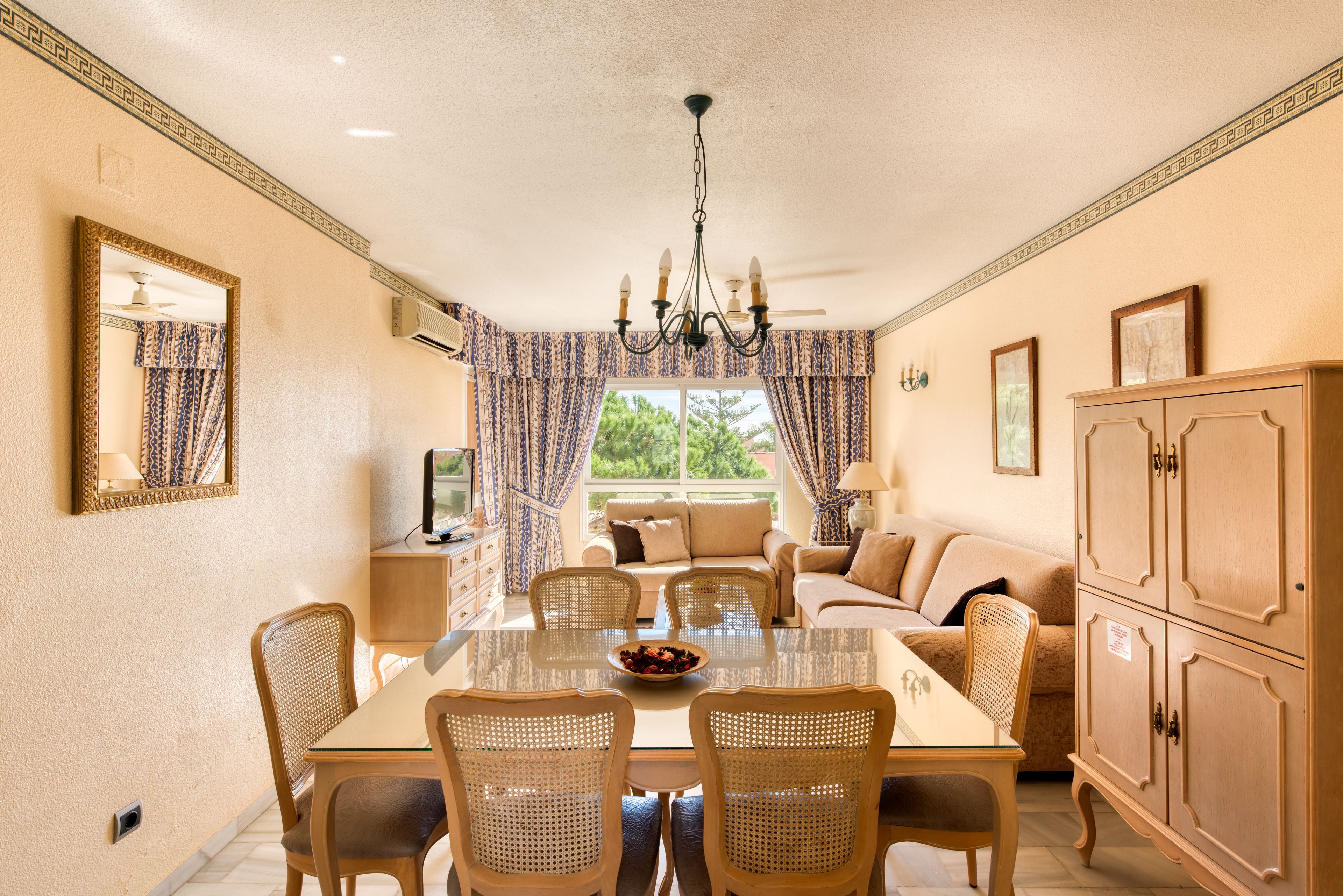 A dining area with a glass table, wicker chairs, a chandelier, and a sofa in the background.