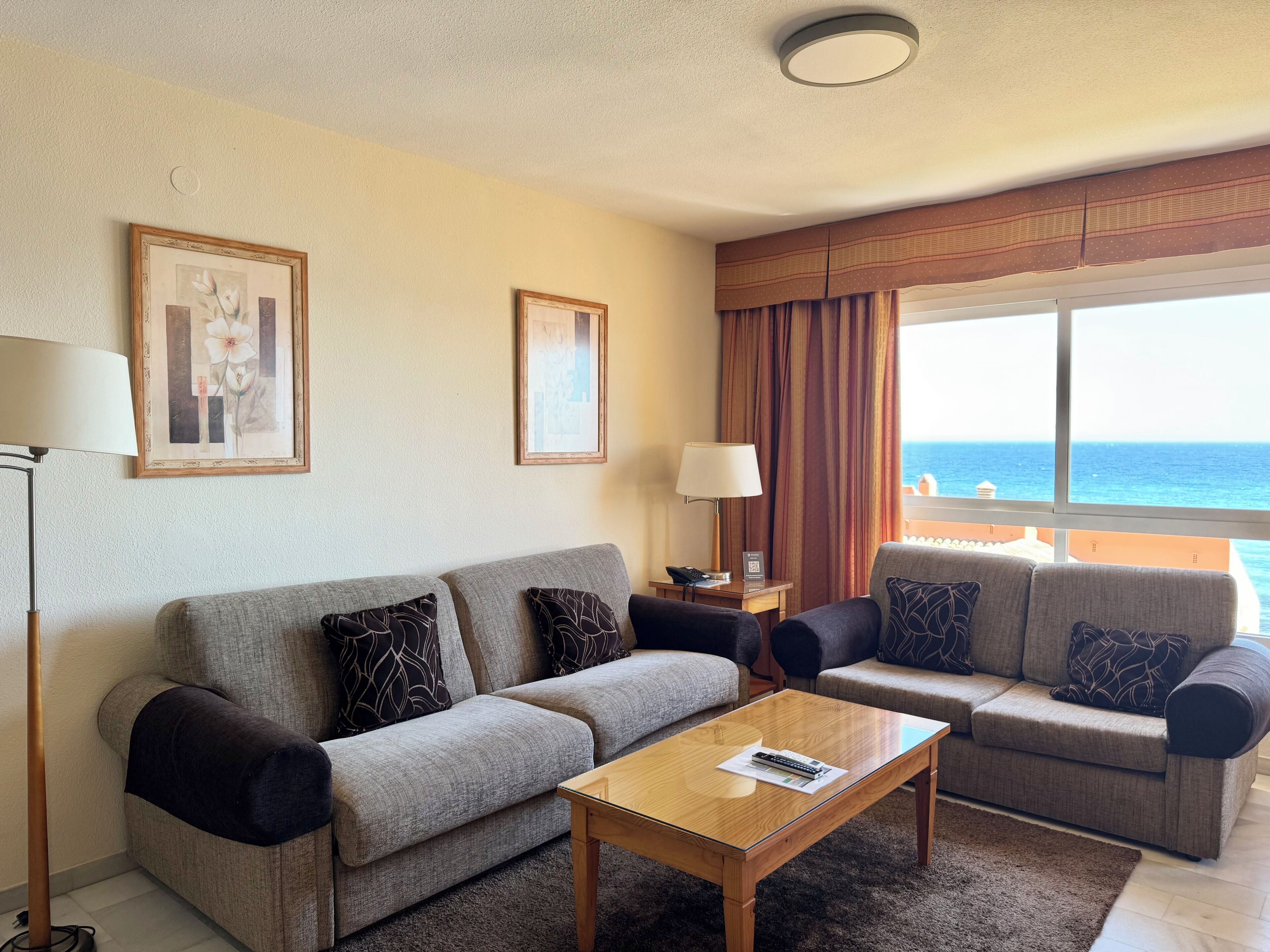 A hotel room with two grey sofas, a wooden coffee table, a floor lamp, and a view of the ocean.