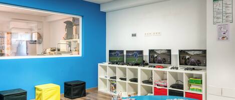 A room with a blue table, a window, and a white wall with shelves containing electronic devices and books.
