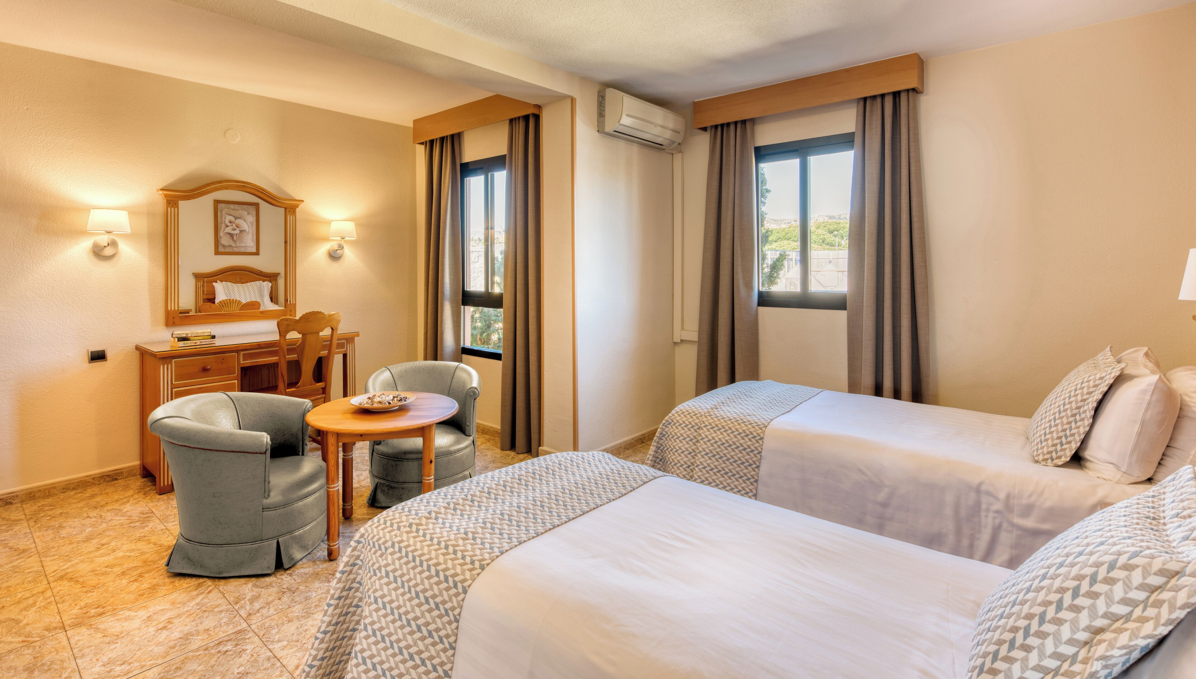 A hotel room with two beds, a desk with a chair, a small table, and a window with curtains.
