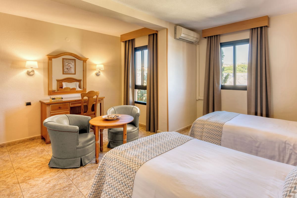 A hotel room with two beds, a desk with a chair, a small table, and a window with curtains.