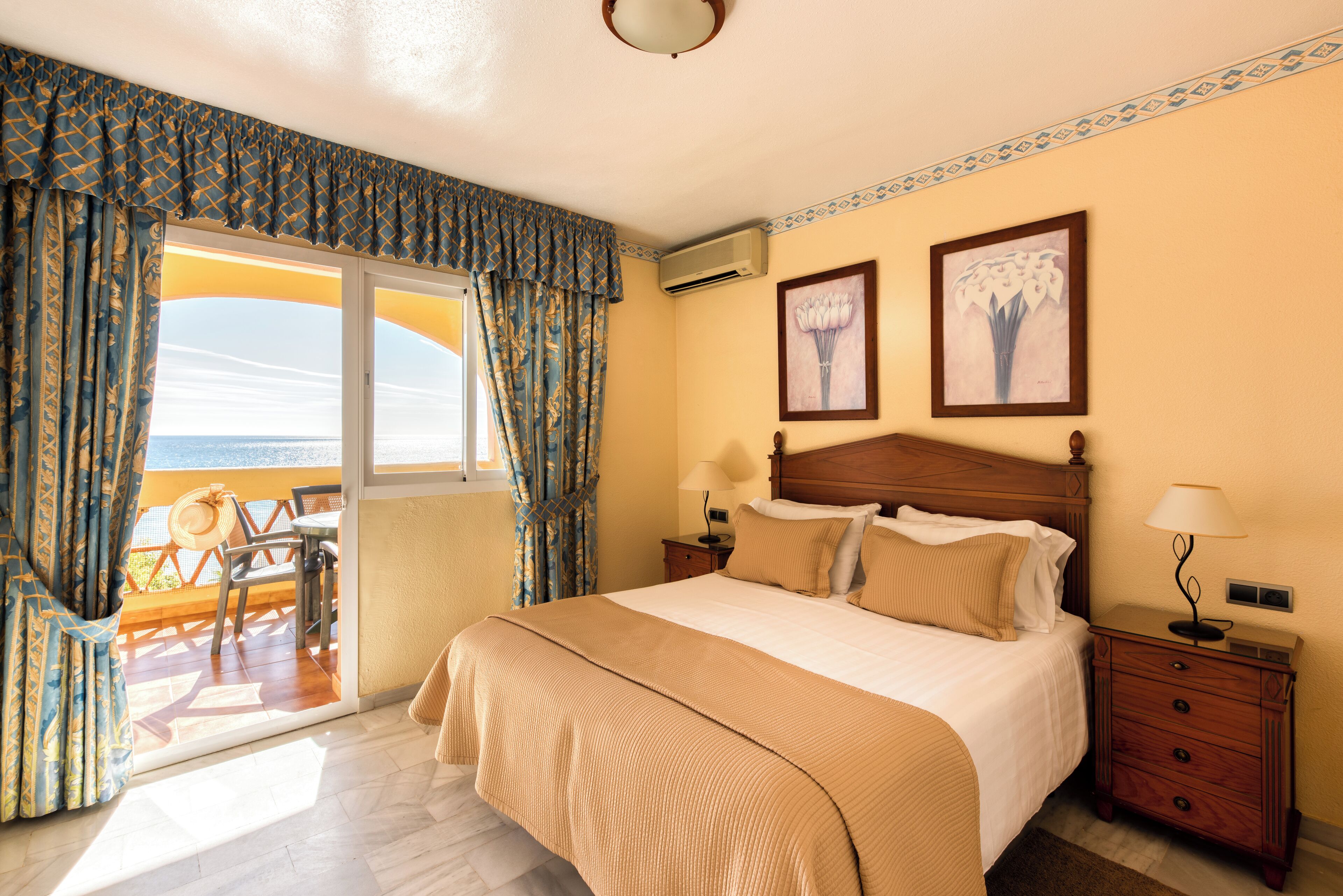Habitación de hotel con una cama grande, una mesita de noche de madera, un escritorio con una silla y vistas al mar a través de un ventanal corredero.