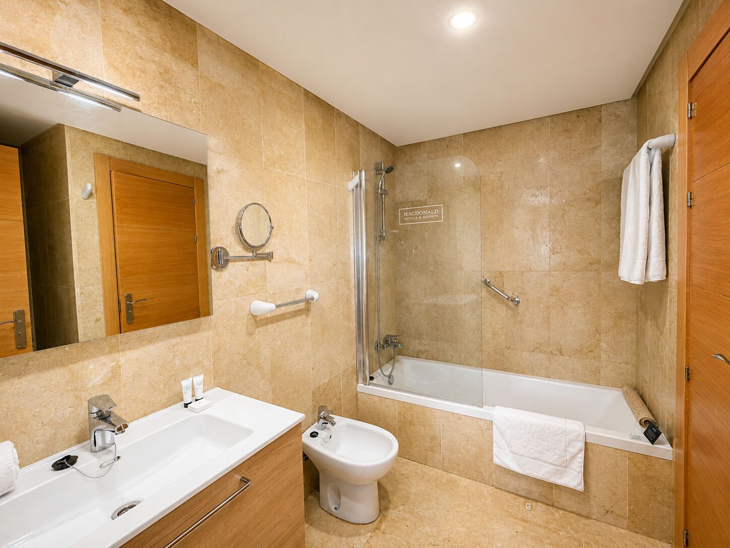 A bathroom with a shower, bidet, and bathtub, wooden cabinets, a mirror, and a sign for Macdonald Hotels & Resorts.