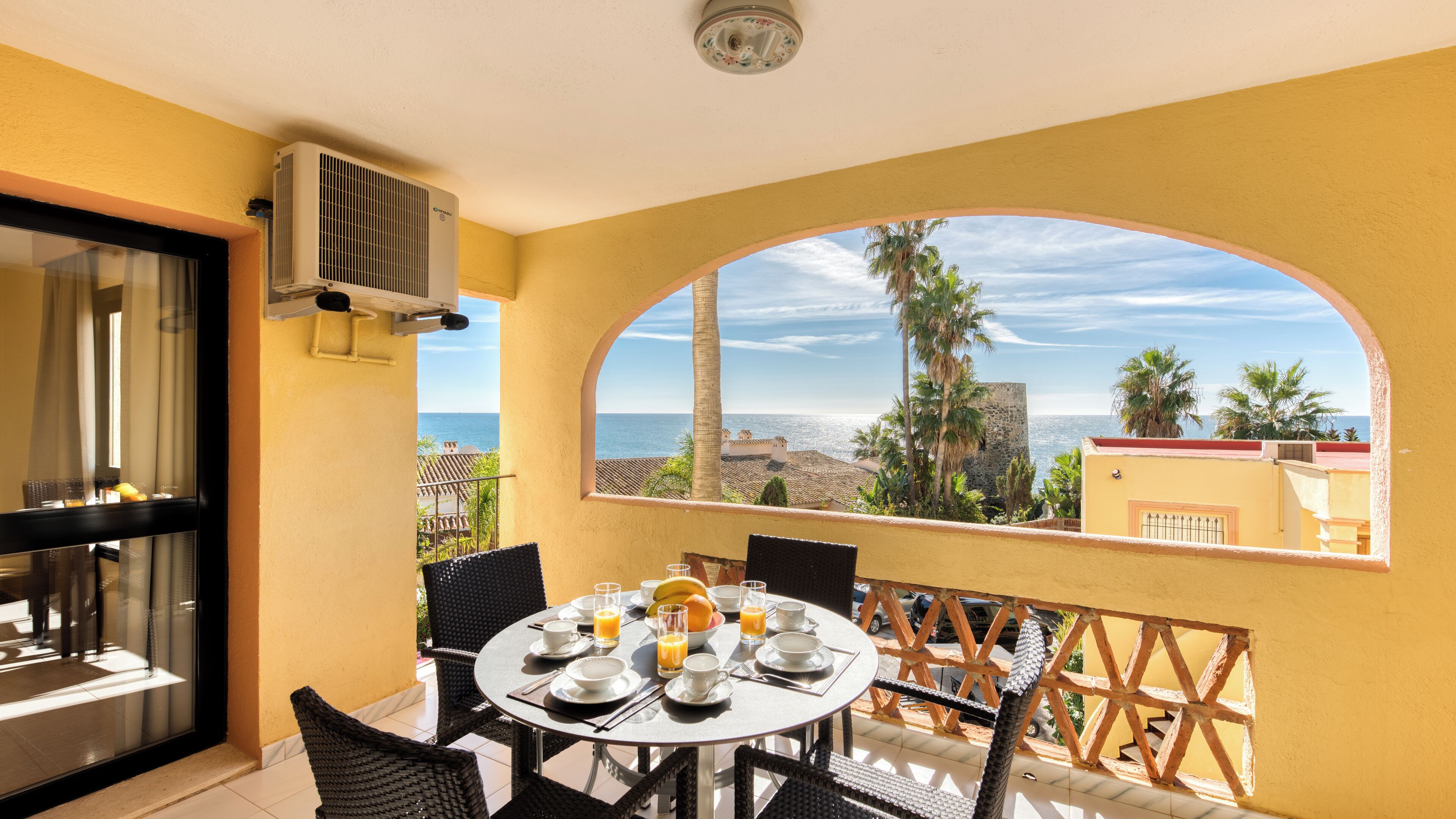 Un balcon avec une table dressée pour le petit-déjeuner, offrant une vue sur la côte, avec des palmiers et des bâtiments.