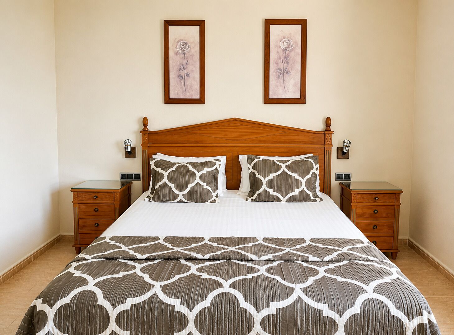 A bedroom with a wooden headboard, two bedside tables, a bed with patterned bedding, and two framed pictures on the wall.