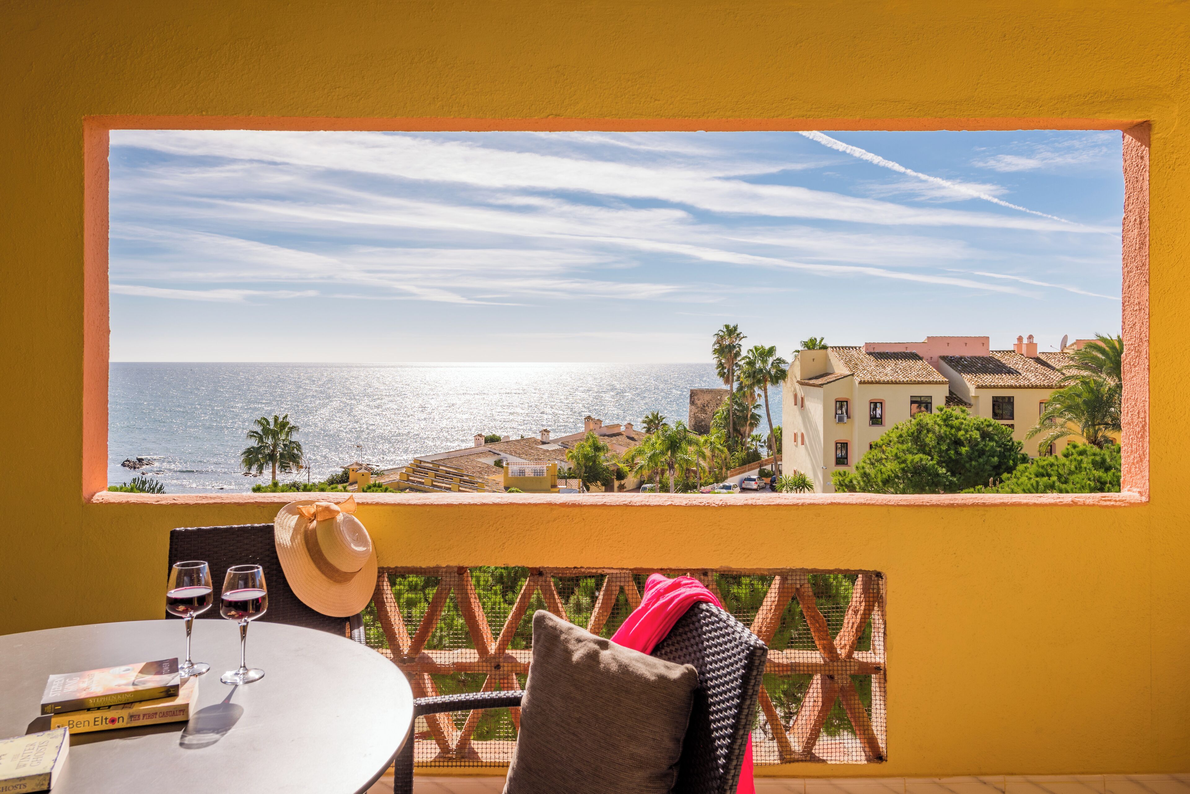 Ein Balkon mit Blick auf das Meer, ein Tisch mit zwei Weingläsern, ein Hut und ein Buch.