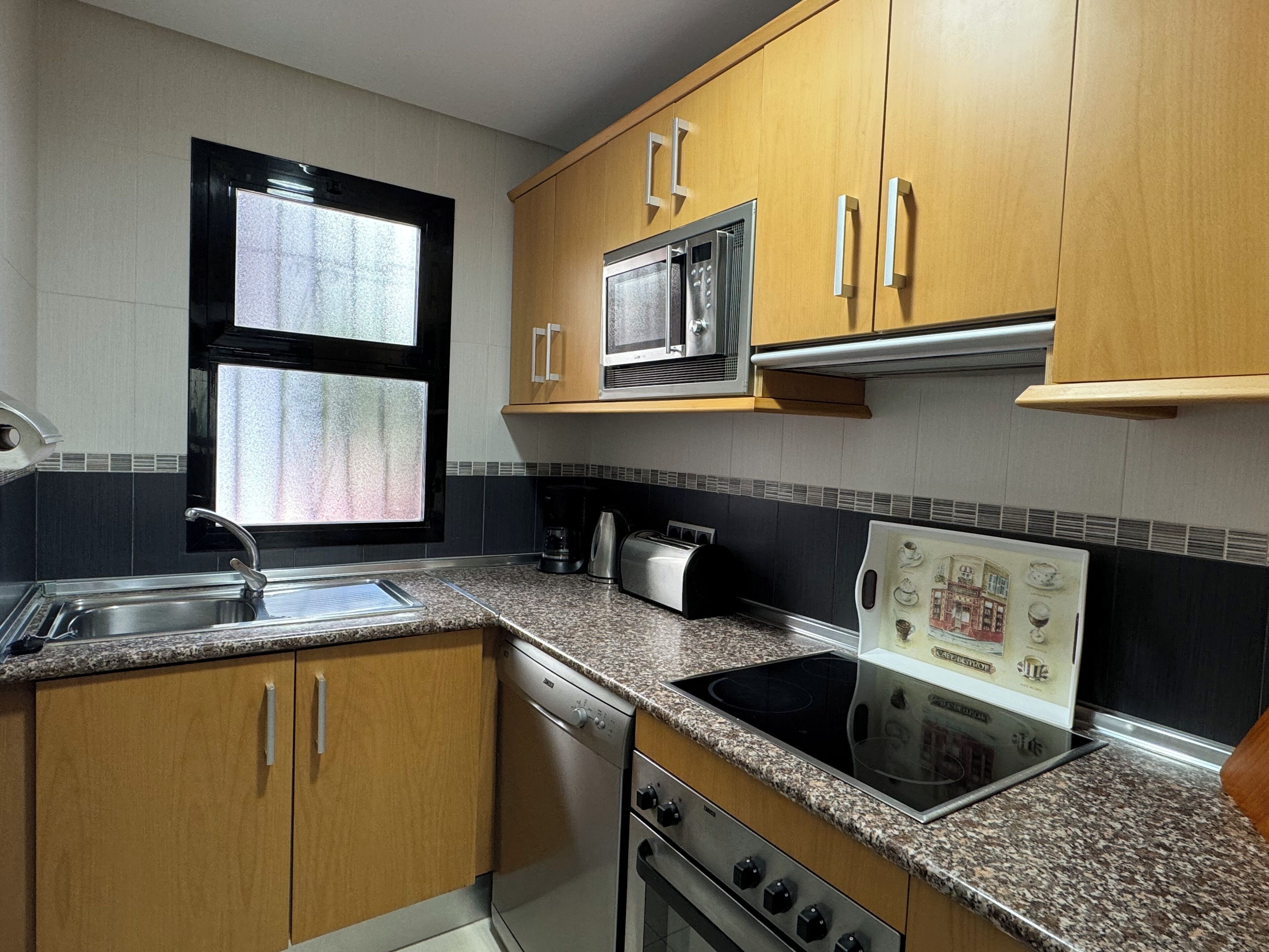 A kitchen with wooden cabinets, a stainless steel microwave, a built-in oven, a stovetop, and a window with frosted glass.