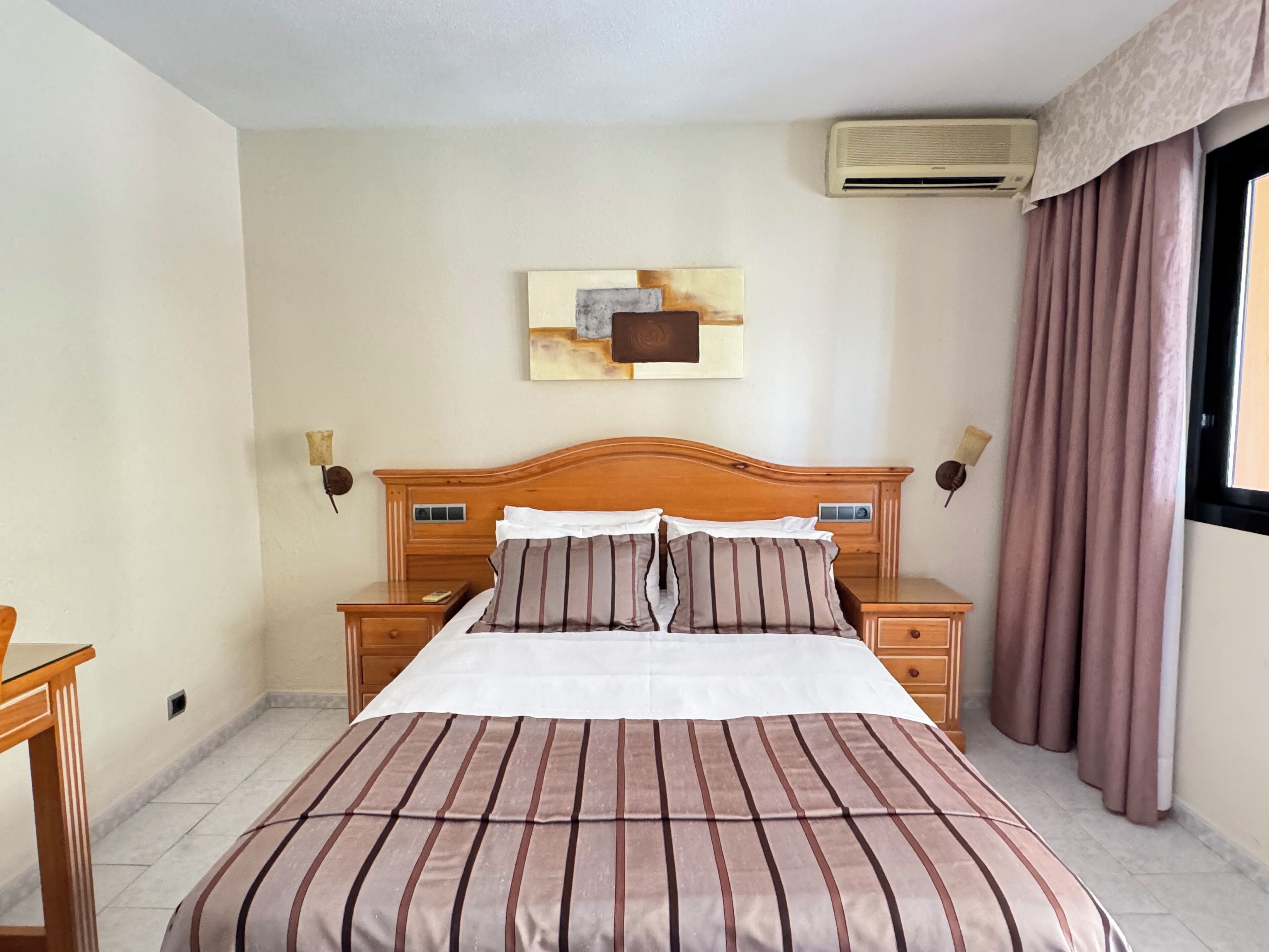 A hotel room with a large bed, wooden headboard, two bedside tables, an air conditioning unit, a window with curtains, and a wall-mounted artwork.