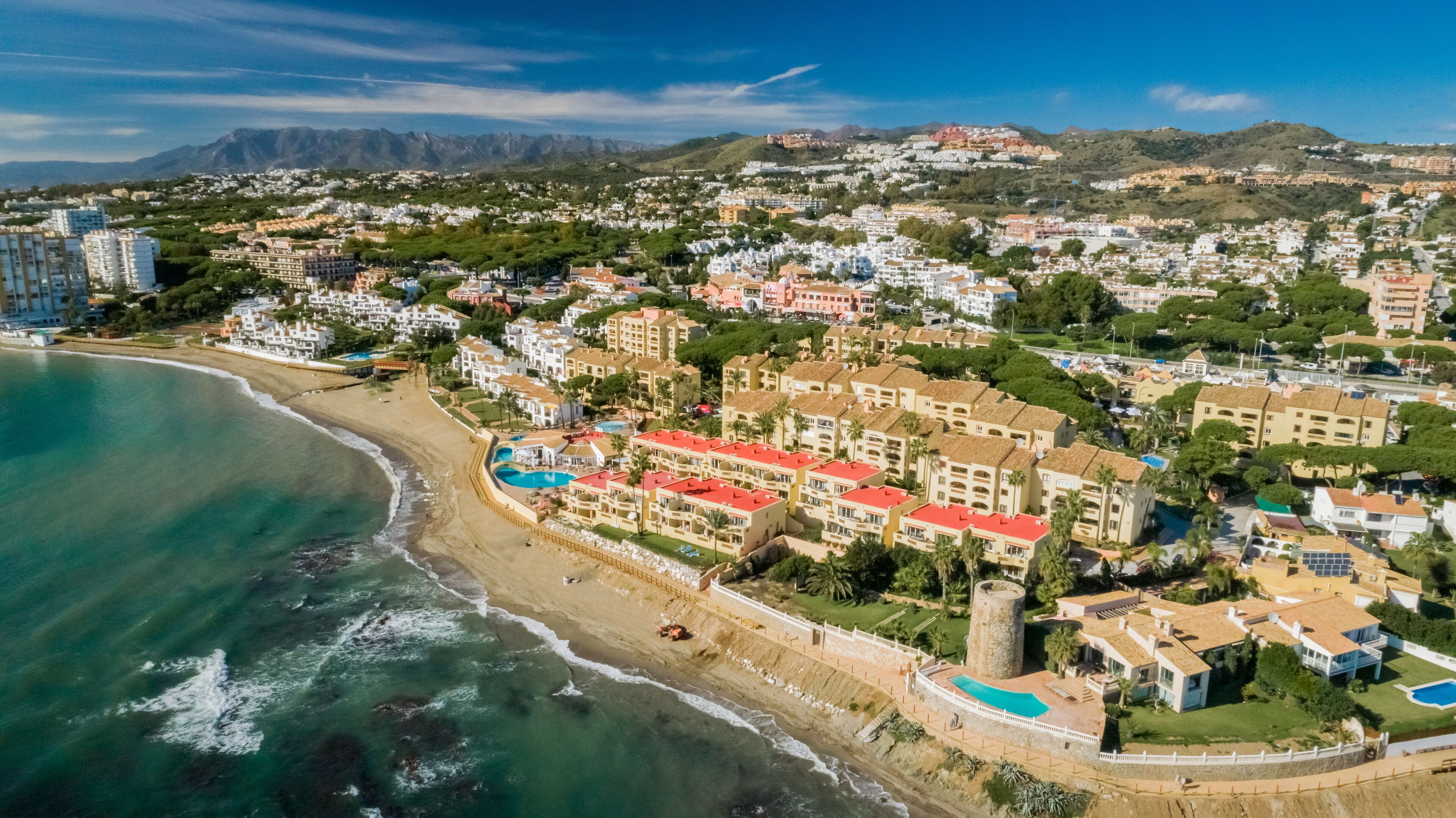 An aerial view of a coastal town with beachfront properties, a large pool complex, and a mountainous backdrop.