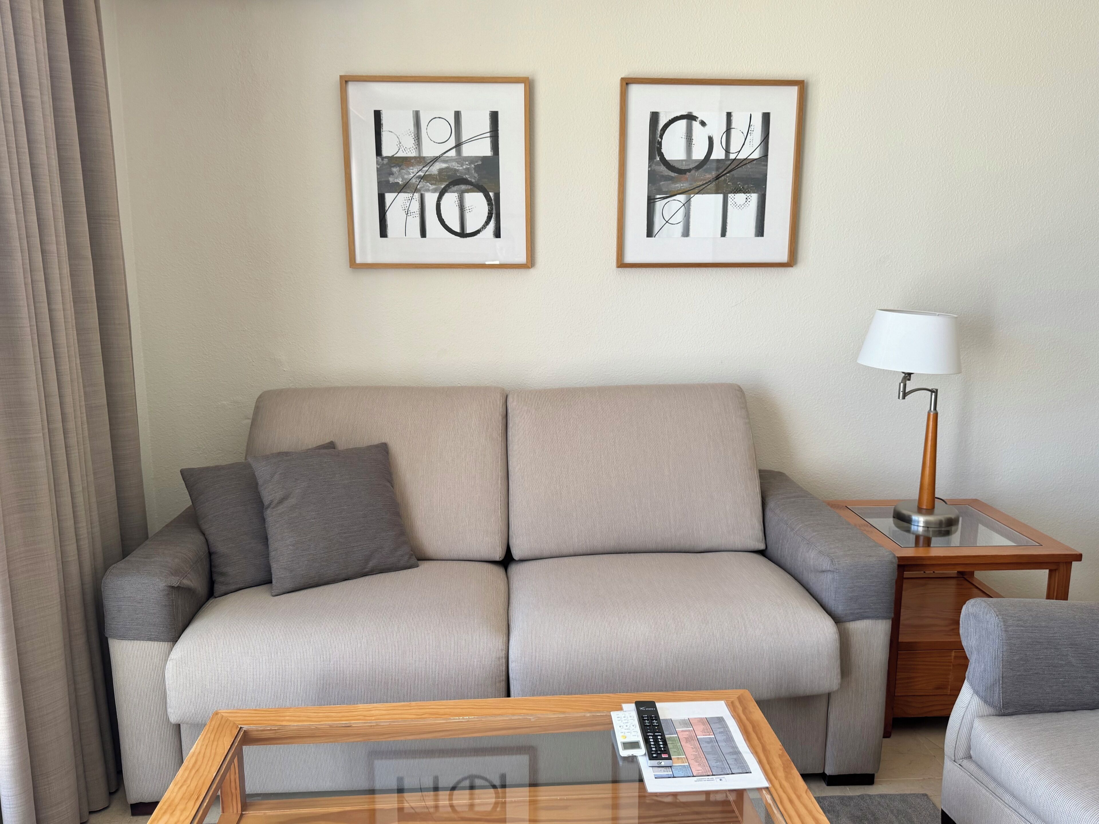 A neutral-toned living room with a sofa, a coffee table with a remote control, and a lamp on a side table. Two framed artworks are hung on the wall.