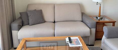 A neutral-toned living room with a sofa, a coffee table with a remote control, and a lamp on a side table. Two framed artworks are hung on the wall.