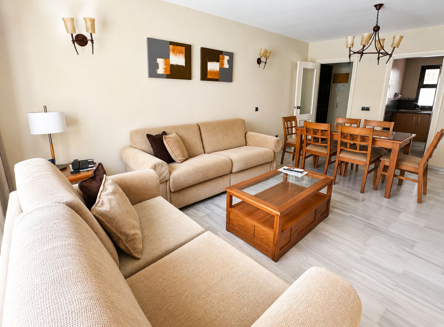 Ein Wohnzimmer mit einer Couch, einem Couchtisch und einem Essbereich mit Stühlen. An den Wänden sind Leuchten angebracht und ein Kronleuchter hängt von der Decke.