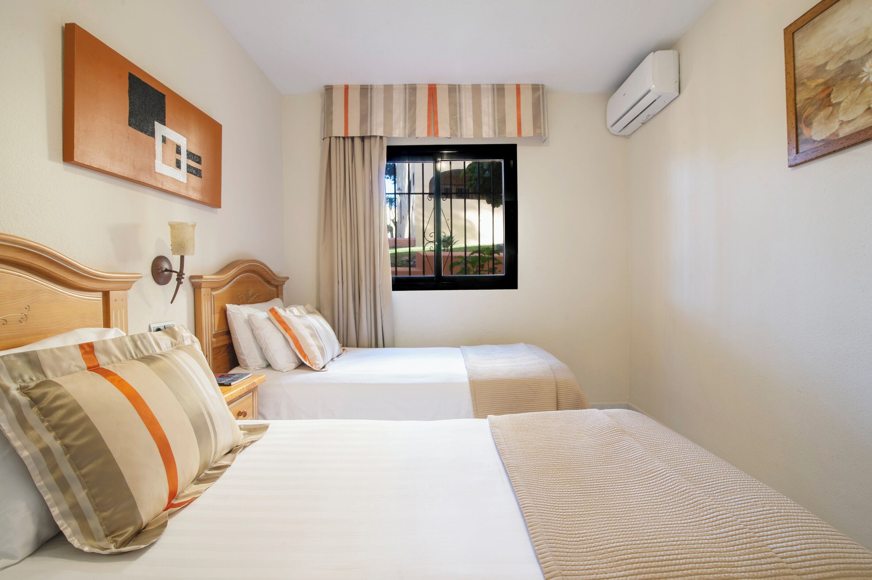 A hotel room with two beds, a window with bars, an air conditioning unit, and a painting on the wall.