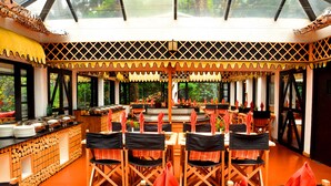 Un restaurante con zona central de comedor que incluye mesas y sillas redondas, un bufé a la izquierda y vistas a zonas verdes a través de amplios ventanales.