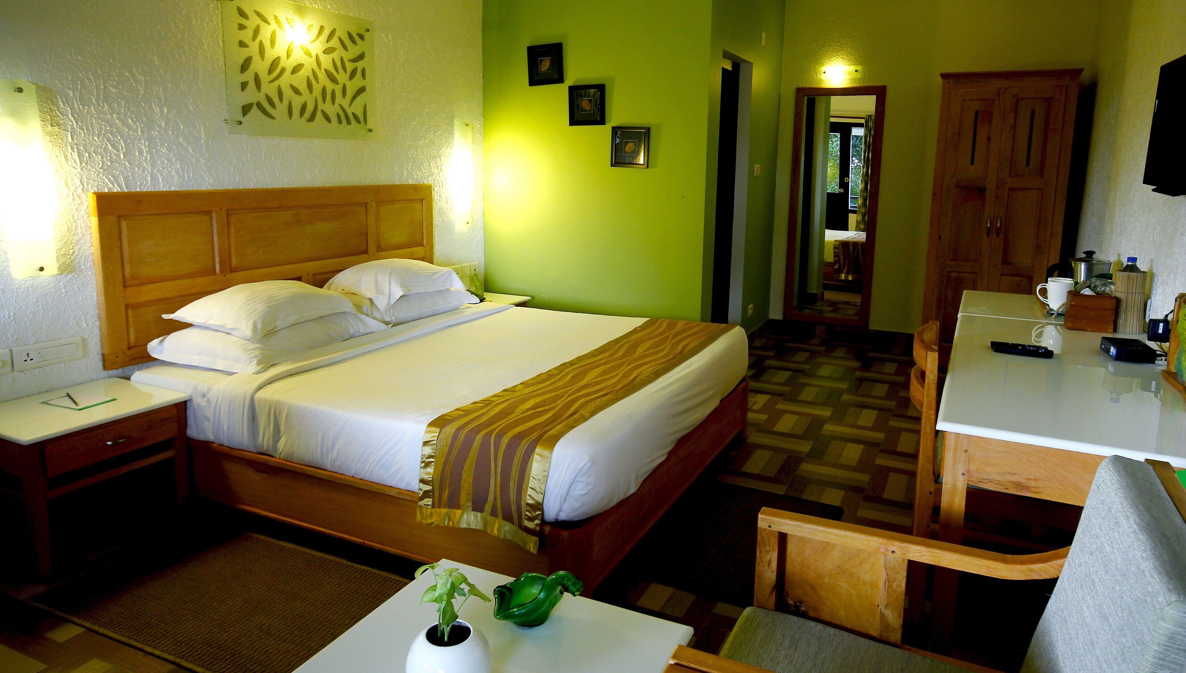 A hotel room with a bed, bedside table, a small table with a plant, a mirror, and a wooden cabinet.