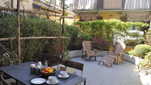 An outdoor patio with a table set for two, surrounded by wooden chairs and a small garden area.