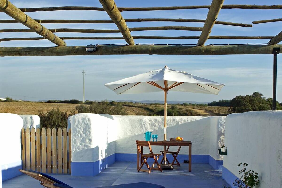 A rooftop terrace with a wooden pergola, a lounge chair, a table with chairs, and a white umbrella.