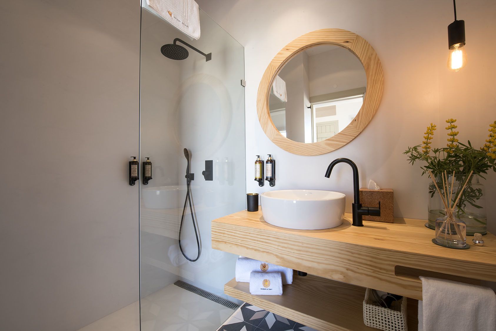Une salle de bain moderne avec une douche à parois de verre, un miroir rond, un meuble-lavabo en bois et un vase rempli de fleurs jaunes.