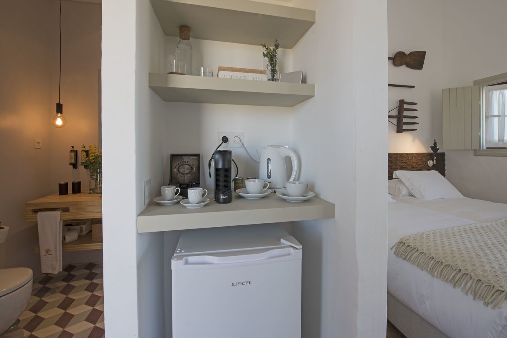 A compact hotel room with a kitchenette, a bed with a headboard, a small refrigerator, and a window with curtains.