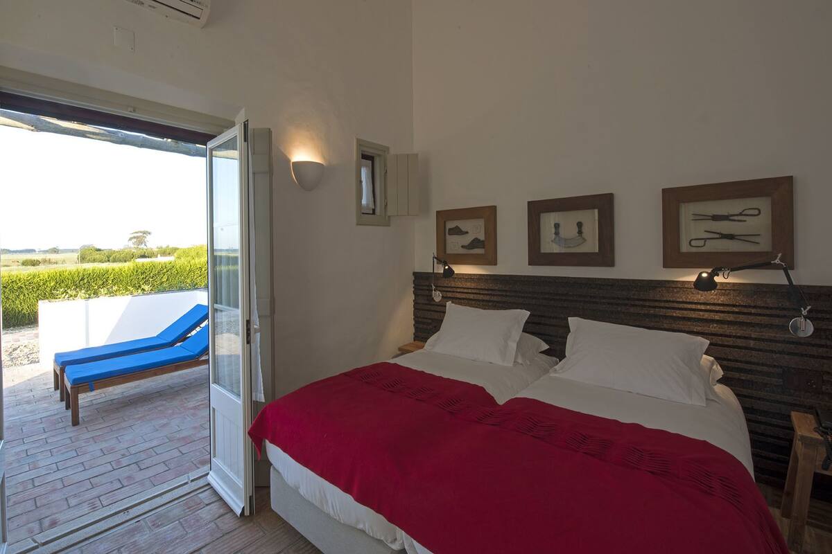 A bedroom with a large bed, a nightstand, and a bedside lamp. There is a window with a view of greenery outside.