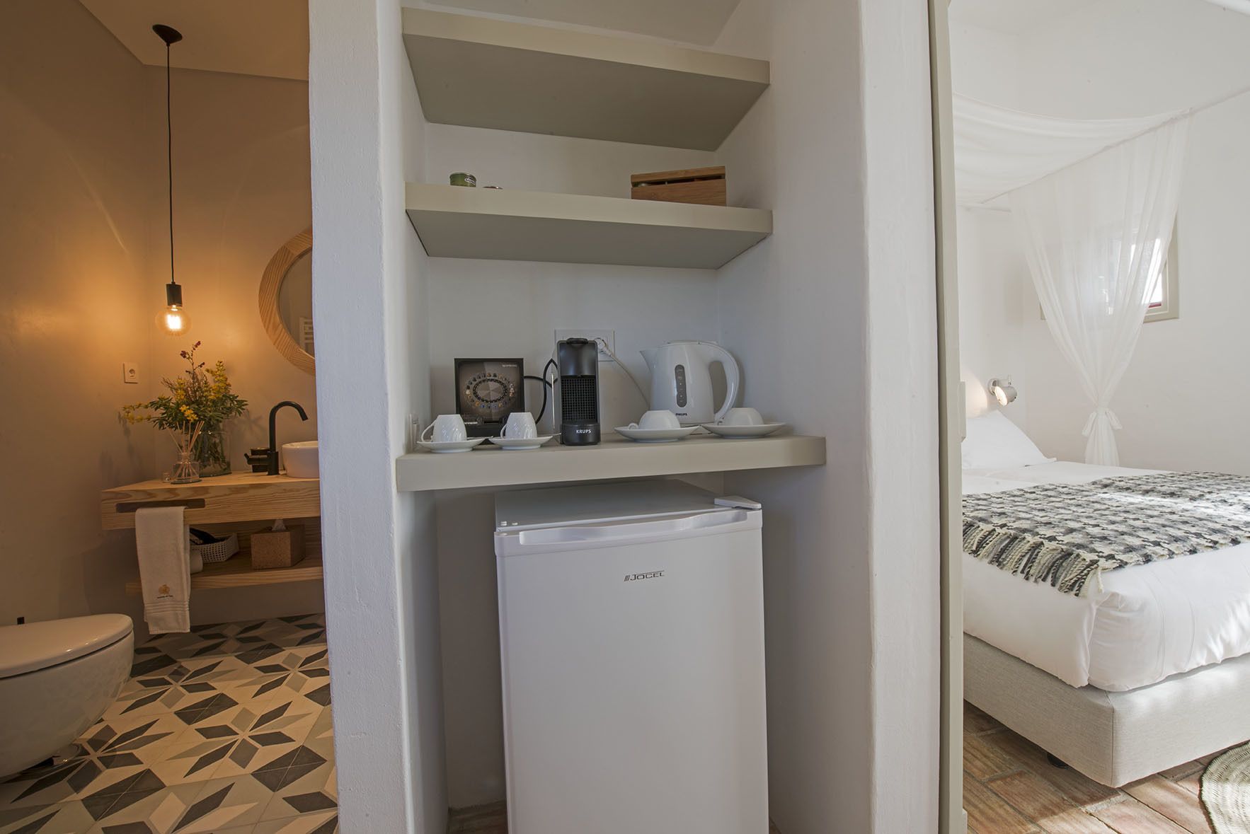 A compact hotel room with a kitchenette, a bed with a canopy, a small refrigerator, and a bathroom with a toilet and sink.