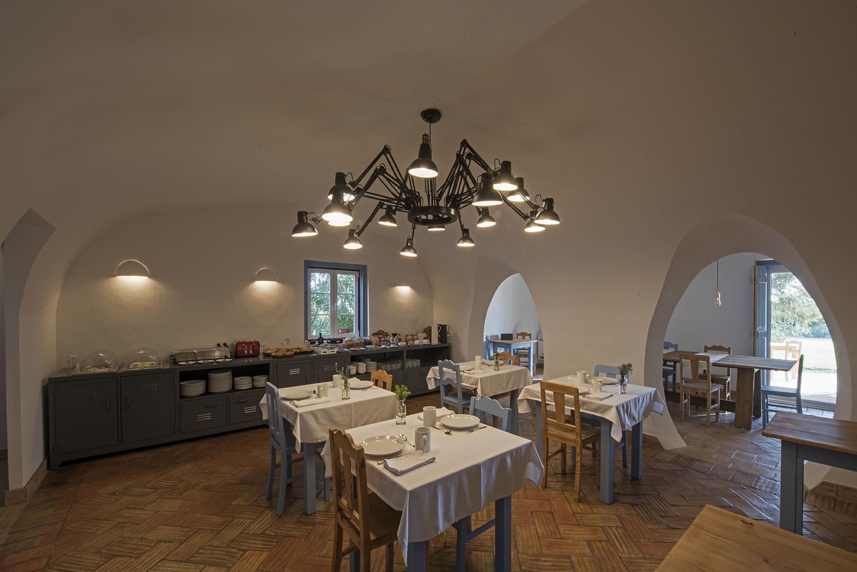 Un vaste espace dédié à la salle à manger, avec de nombreuses tables et chaises, un lustre central et un coin cuisine équipé d’un buffet.