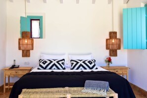 A bedroom with a bed, two bedside tables, a nightstand, a woven bench, and a hanging light fixture.