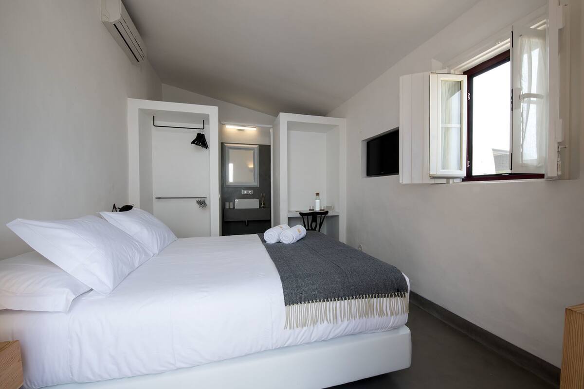 A modern hotel room with a large bed, a small desk, a mirror, and a window with curtains.