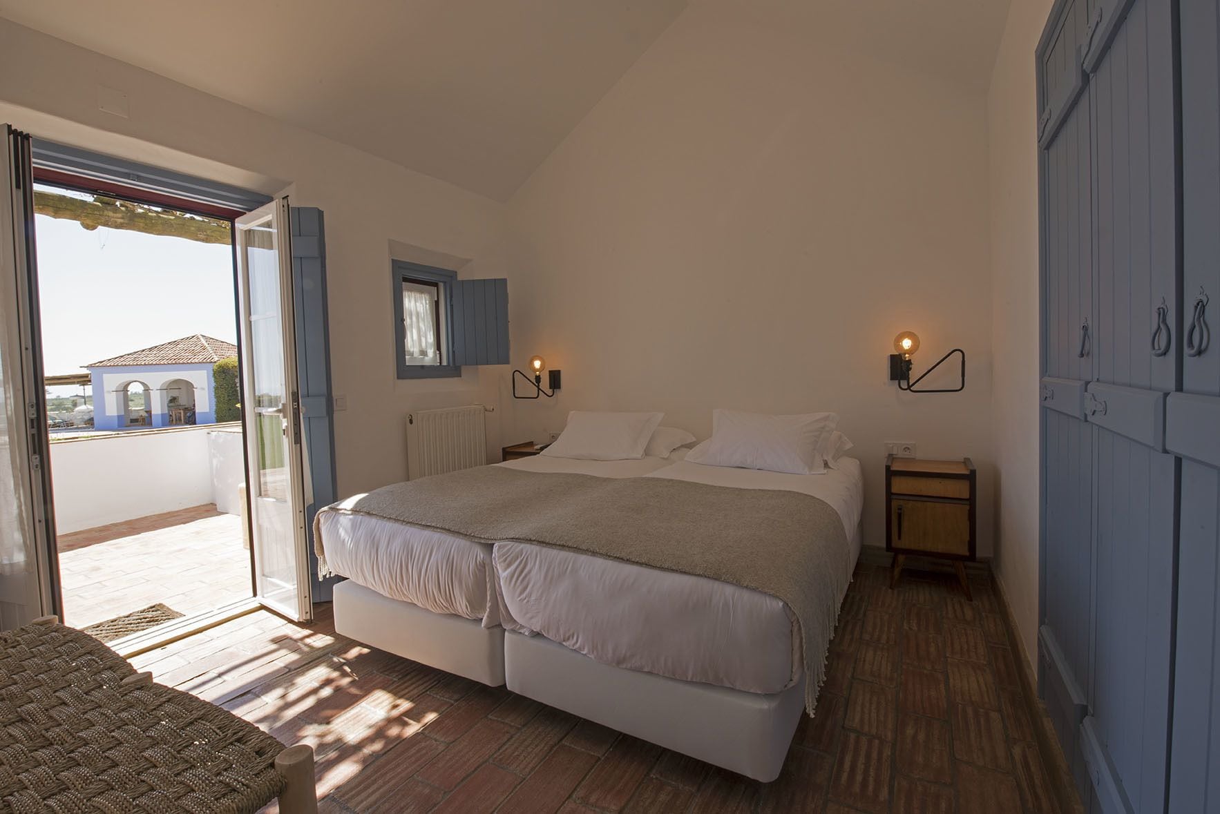 Une chambre avec un grand lit, une table de chevet, deux lampes fixées au mur et une vue sur un bâtiment à l’extérieur.
