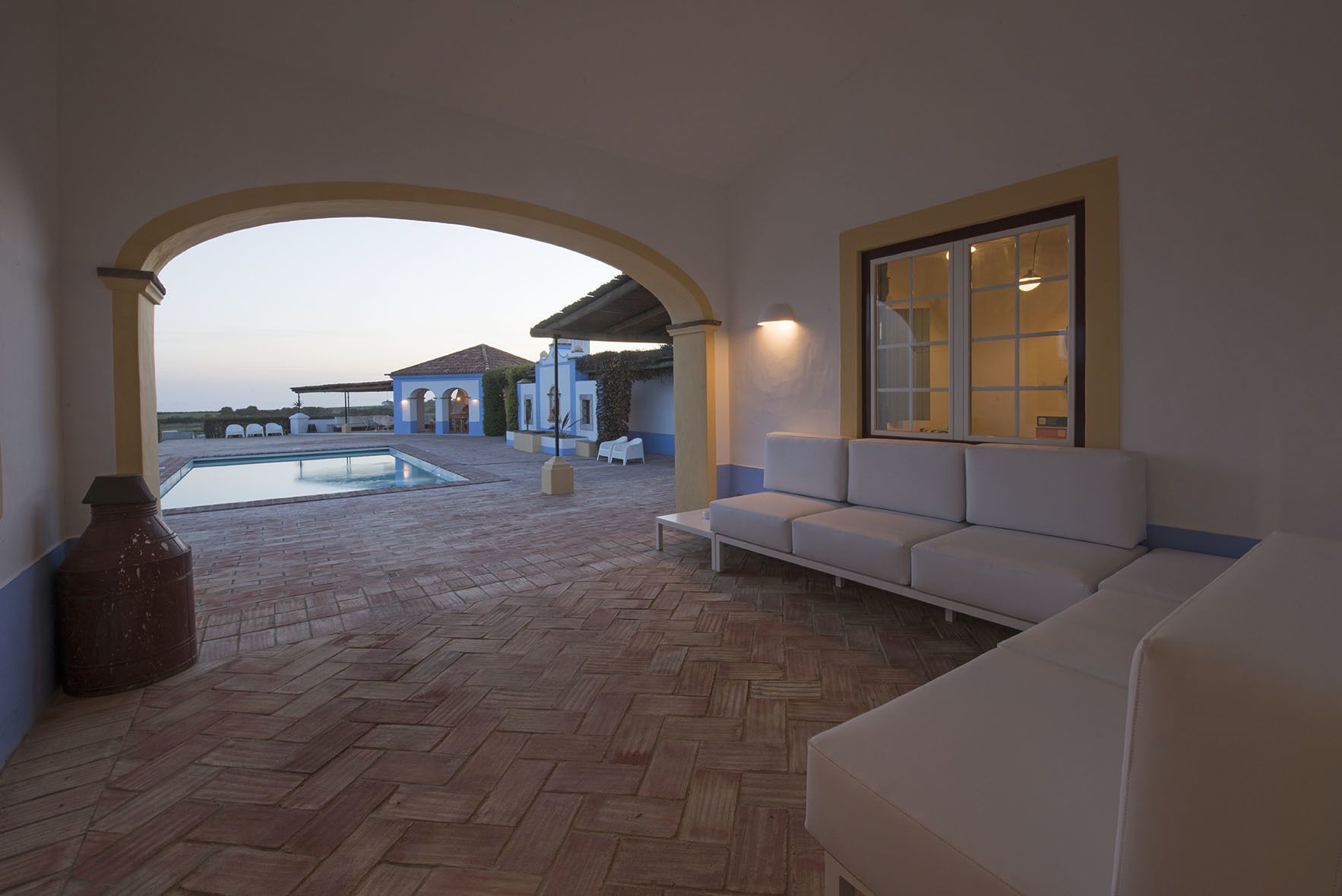 Un espacio exterior techado con piscina, un sofá blanco y un ventanal grande.