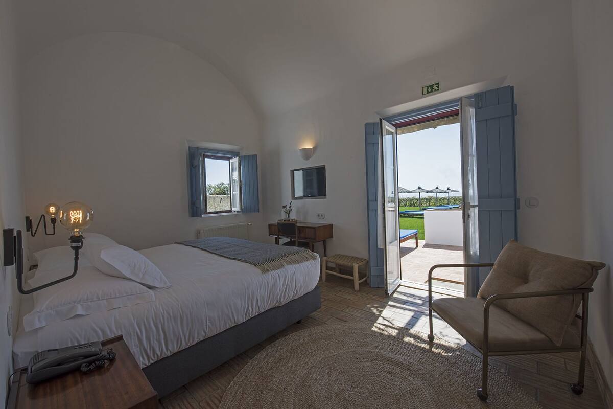 A bedroom with a bed, bedside lamp, desk, chair, and an open door to a balcony.