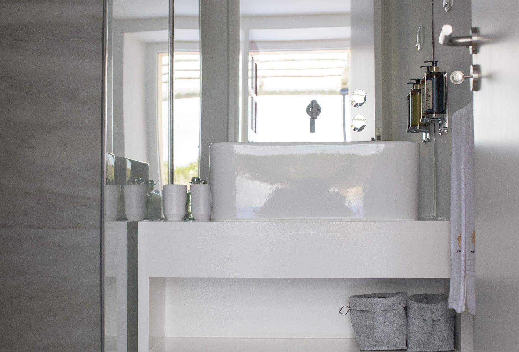 Un baño moderno con un lavamanos grande blanco, un espejo y una ventana. Hay artículos de aseo en el lavamanos y un perchero con toallas.