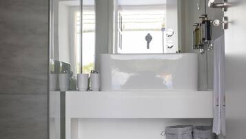 Un baño moderno con un lavamanos grande blanco, un espejo y una ventana. Hay artículos de aseo en el lavamanos y un perchero con toallas.