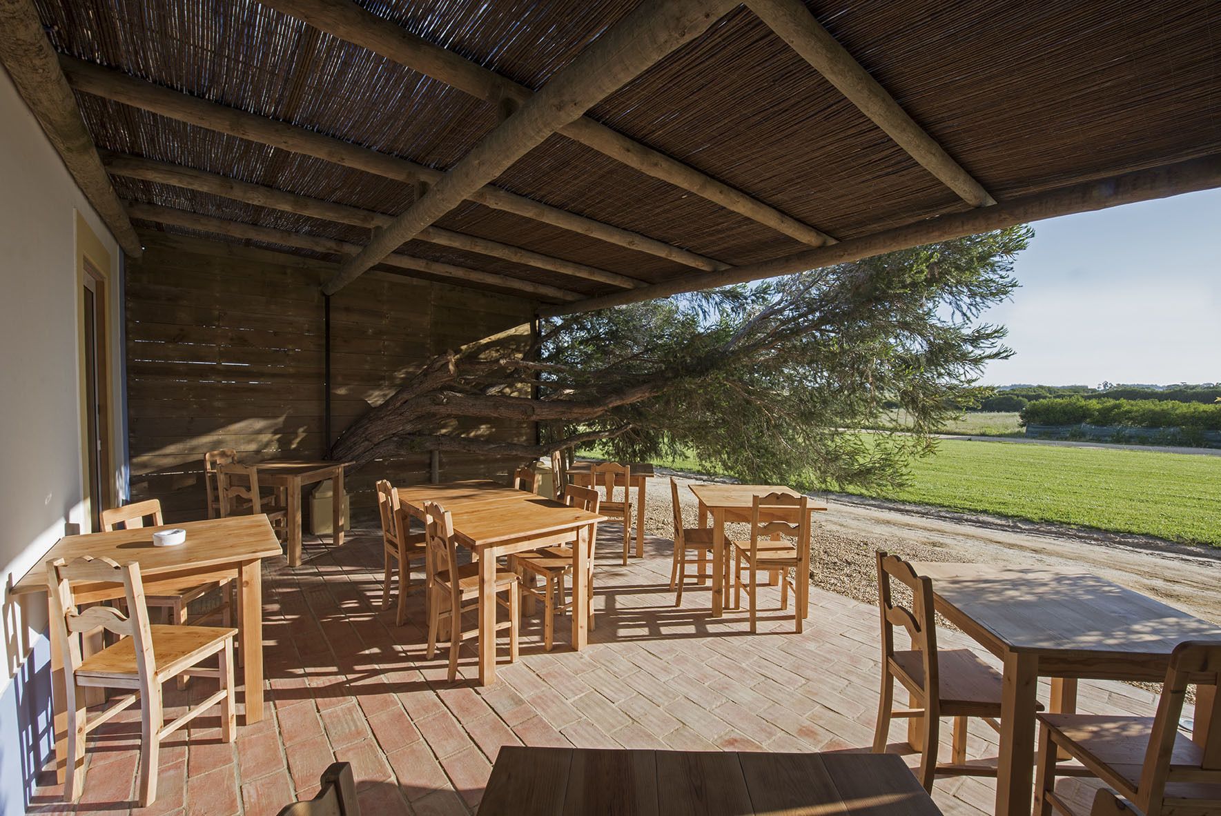 Une terrasse extérieure en bois, dotée de tables et de chaises, d’un toit de chaume et offrant une vue sur un champ verdoyant.