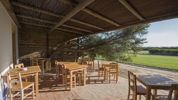 Une terrasse extérieure en bois, dotée de tables et de chaises, d’un toit de chaume et offrant une vue sur un champ verdoyant.