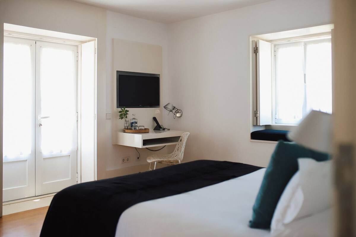 A bedroom with a bed, a desk with a computer, a TV, and a window.