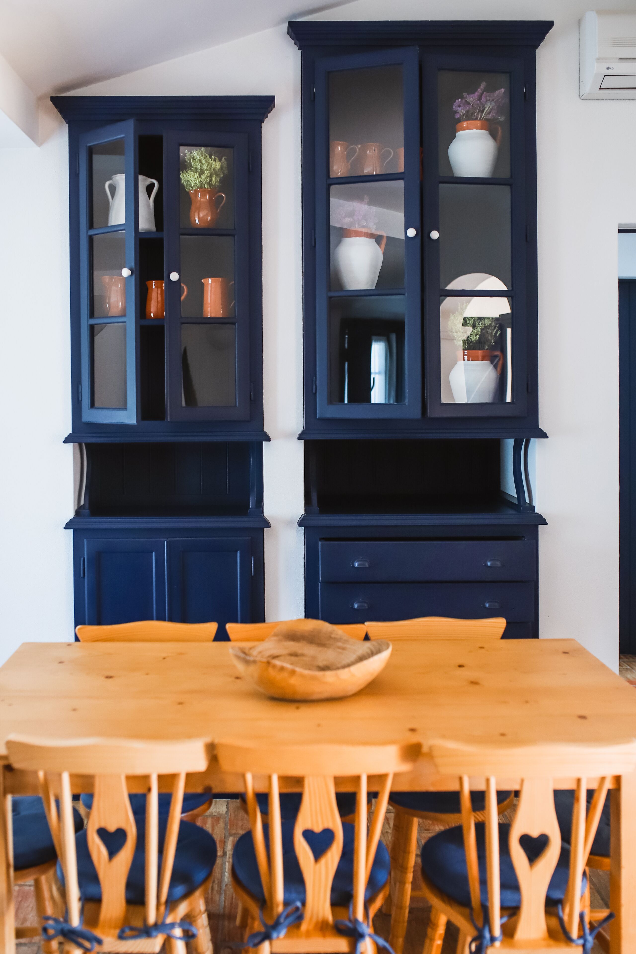 A dining area with a wooden table and chairs, a dark blue cabinet with glass doors, and a white air conditioning unit on the wall.